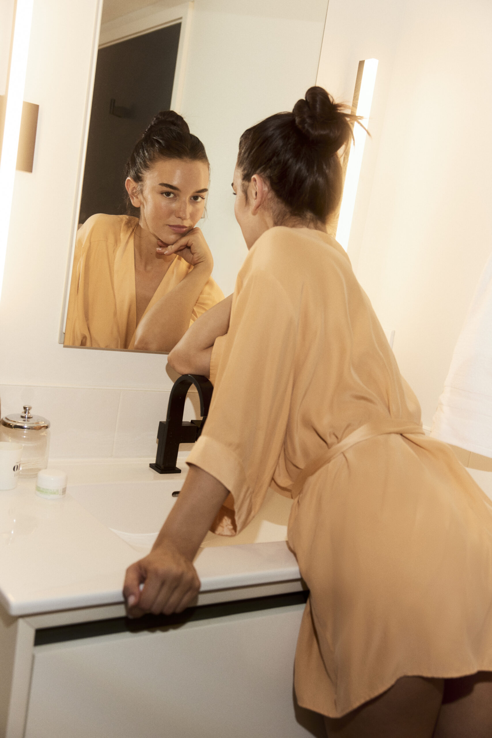 A young woman wearing a bathrobe looking at the camera through the mirror in front of her.